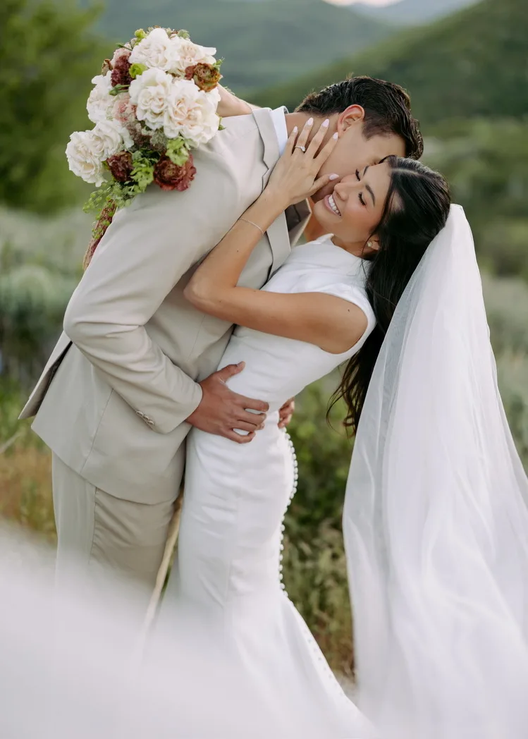 A groom holds his bride close outdoors against a green hillside, her veil falling behind her and a cream and burgundy bouquet resting over his shoulder.