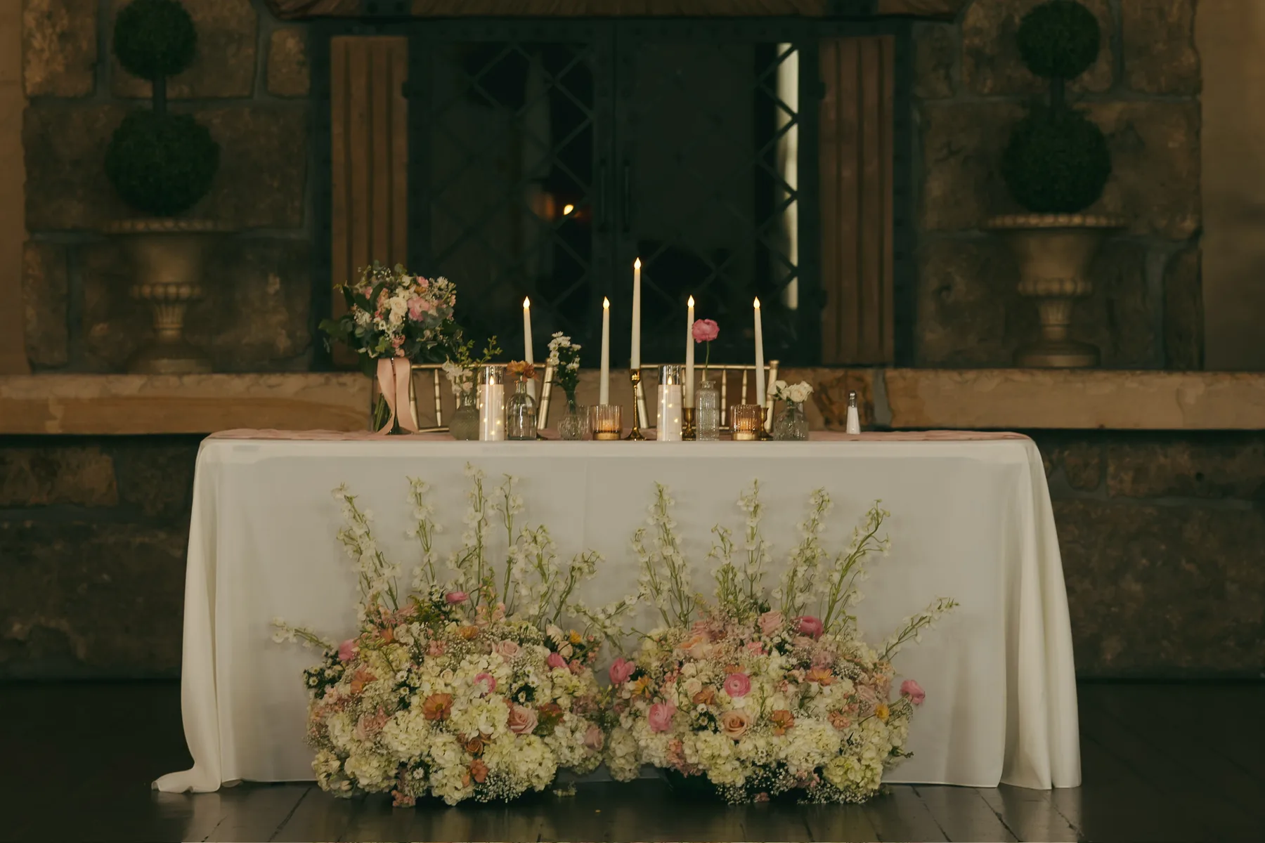 A long wedding head table under a garland of white and blush flowers and trailing greenery, with tapered candles in glass holders down its length.